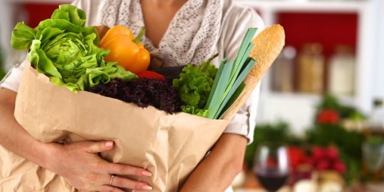 43218252 - young woman holding grocery shopping bag with vegetables standing in the kitchen.
